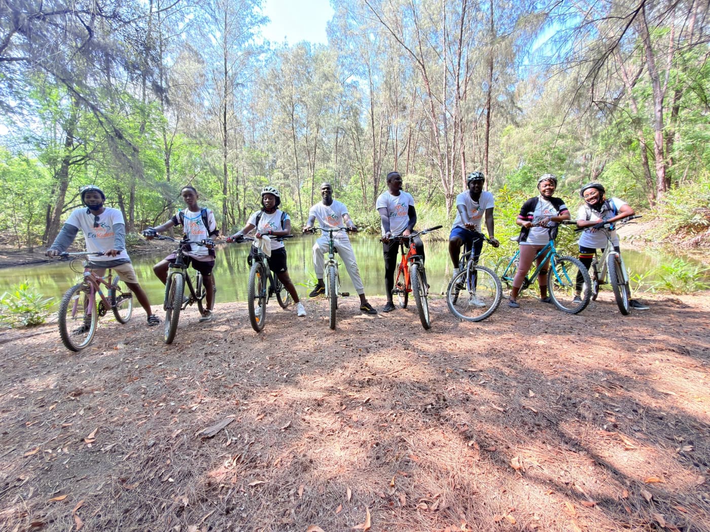 A group of riders with their bikes pausing by a green creek lined with trees