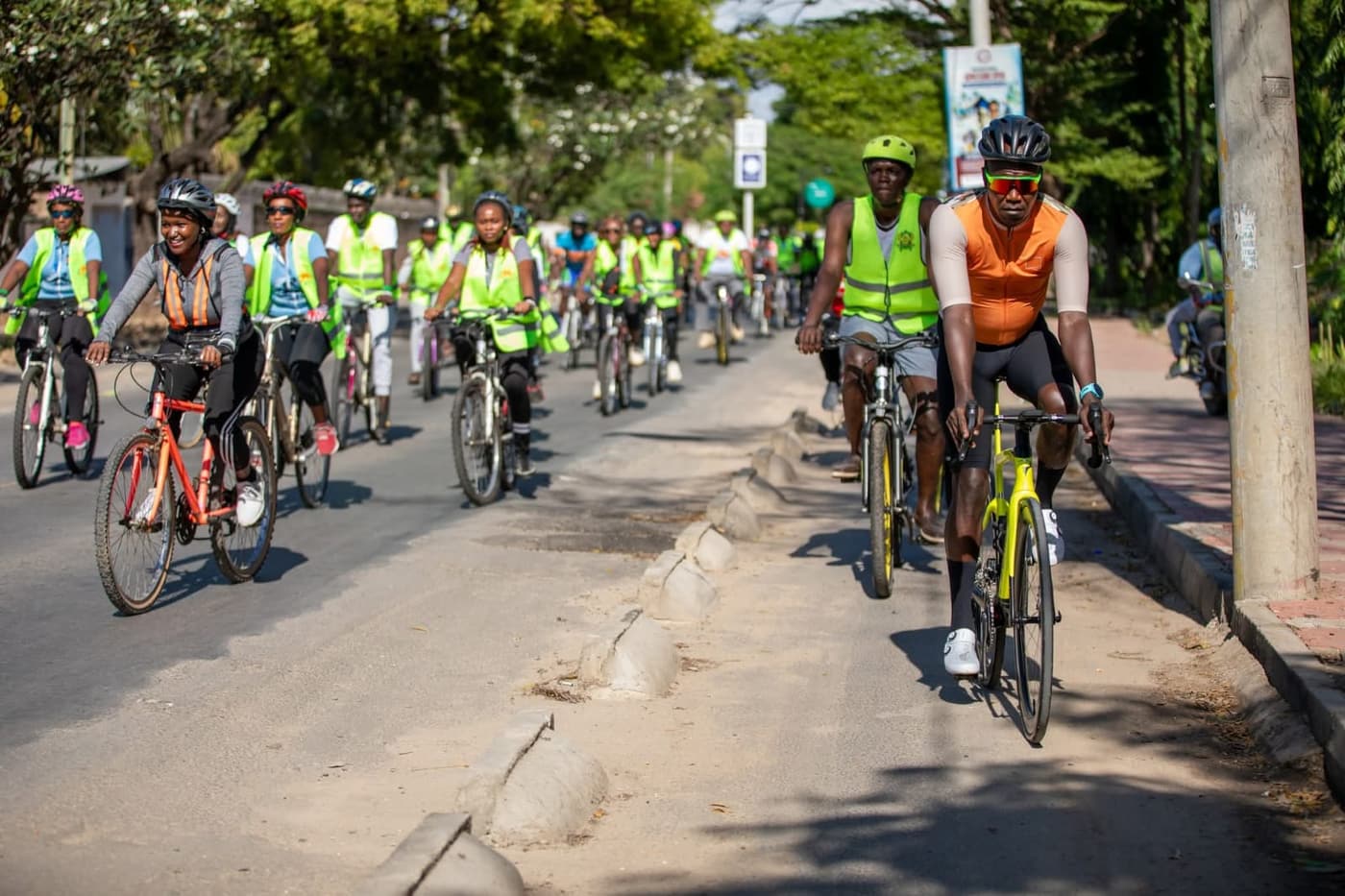 A column of riders in hi-vis vests on a Mombasa road, led by a cyclist in orange kit