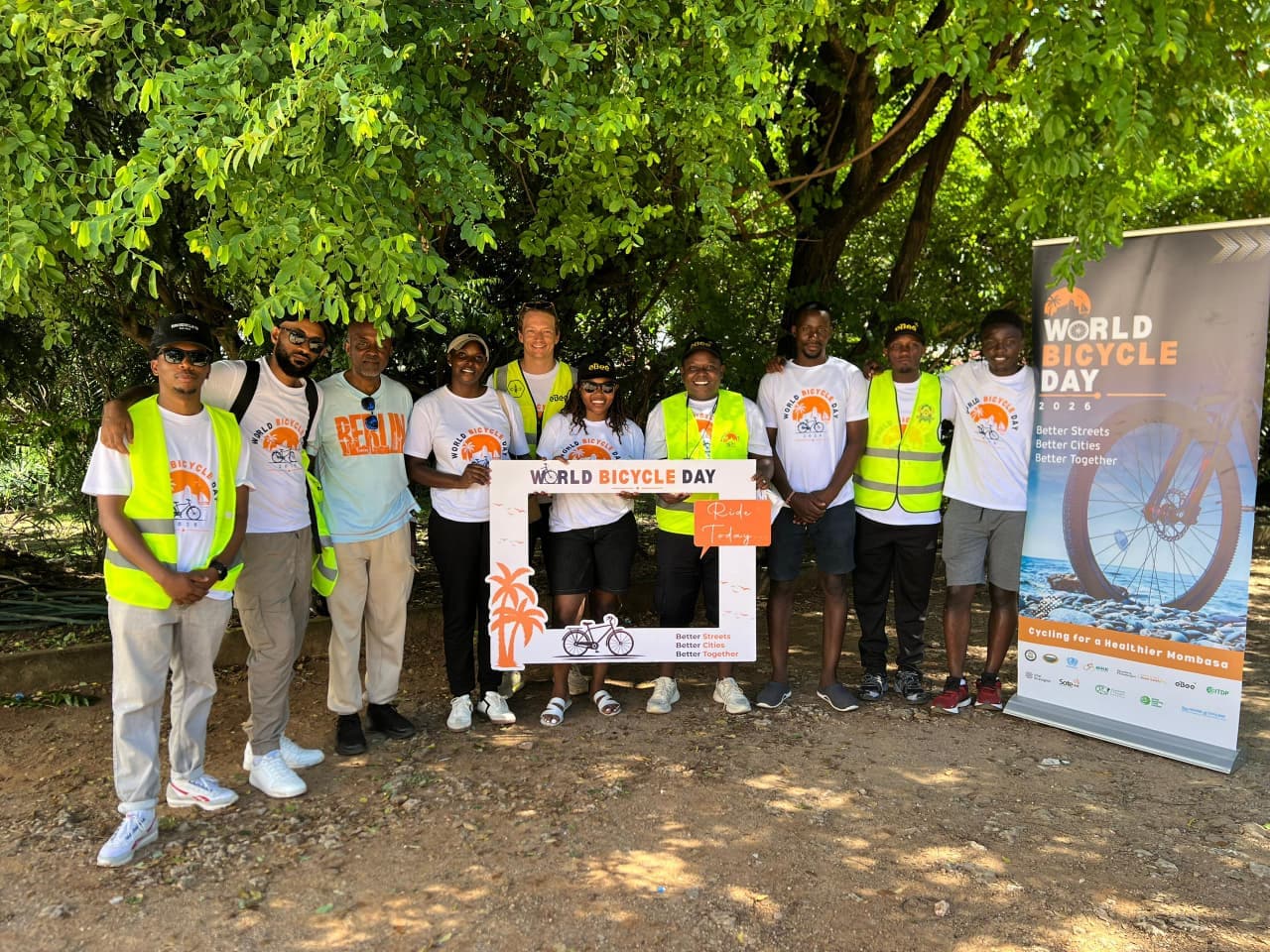 The Bike is Best team under a tree holding a World Bicycle Day board beside a 'Cycling for a Healthier Mombasa' banner